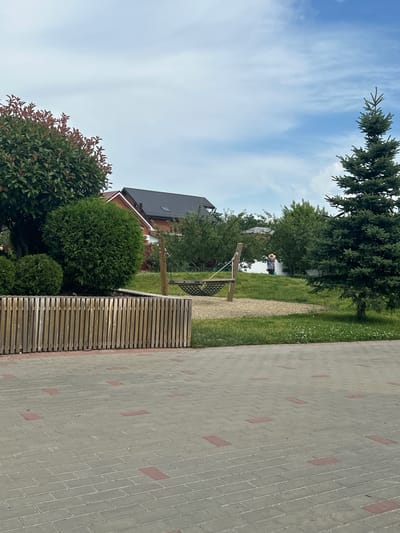 This image depicts an outdoor scene with a paved walkway in the foreground and a grassy area with trees and buildings in the background. The walkway is made of grey interlocking pavers with occasional red rectangular inserts. A wooden fence, constructed from vertical planks, runs horizontally across the middle ground. Beyond the fence, a grassy slope features a hammock suspended between two wooden posts. A large evergreen tree stands to the right. In the background, several houses with dark roofs are visible, nestled amongst trees. A person wearing a hat and a light-colored top is walking on the grassy slope in the distance. The sky is partly cloudy with blue patches. The overall setting appears to be a residential or park area.