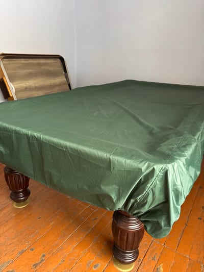 The scene depicts an indoor environment in Bayronovka, Russia. A large rectangular table, likely a billiard table, is positioned prominently in the foreground and midground, covered entirely by a dark green, slightly wrinkled protective cover. The cover drapes over the edges and covers the upper portions of the table's four visible, turned wooden legs, which rest on circular, light-colored floor protectors. The floor consists of orange-brown wooden planks exhibiting wear, chipping, and scratches. In the background, leaning against a plain white wall in the corner of the room, is a light brown wooden tray with a textured, possibly laminate, surface. No people or active interactions are present.