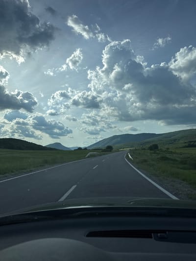 The image depicts a road in Medven, Bulgaria. The road is asphalt with white line markings, flanked by green grassy areas and a line of trees on the left. A mountain range is visible in the distance on the left. The sky above is filled with numerous white clouds and some patches of blue sky. The image is taken from inside a vehicle, with part of the interior visible on the right side.