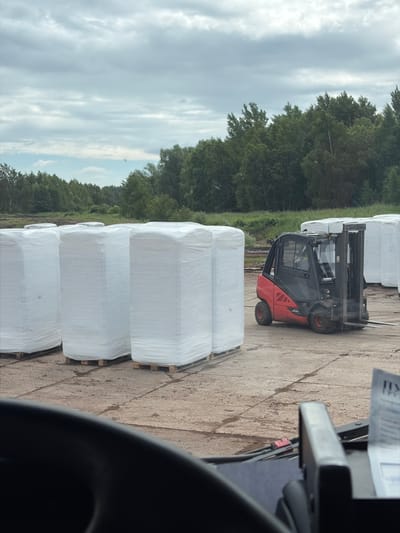 An exterior shot captures a cloudy day in Zarech'e, Russia. In the foreground, a dark vehicle dashboard is visible, with a piece of paper displaying Russian text in the lower right corner. The middle ground features a red and black forklift with its forks extended, positioned on a concrete surface. To the left of the forklift, several large, rectangular white bales wrapped in plastic are stacked on wooden pallets. In the background, a dense line of green trees stretches across the horizon, under a sky filled with gray and white clouds.