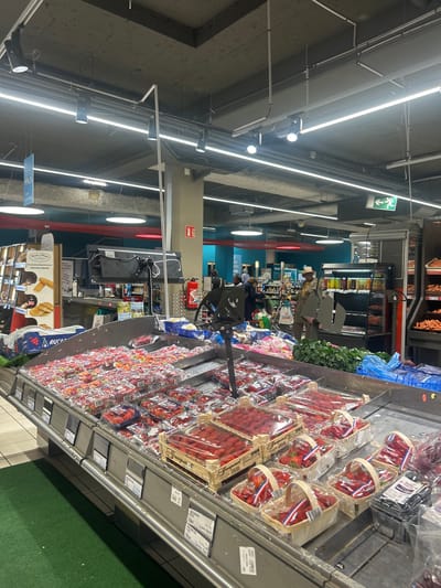 This image captures an indoor scene within a modern supermarket or grocery store, likely in Noisy-le-Grand, France, given the visible French text. The primary focus is a large produce display, predominantly featuring numerous packaged strawberries.
The scene is brightly lit by linear fluorescent lights and spotlights mounted on the high, concrete ceiling, which also features exposed pipes and wiring. In the foreground, dozens of plastic containers and wooden crates filled with strawberries are meticulously arranged on a low display unit. Some containers bear labels such as "Dream Manann's" and "Guapert FRANCE". Green artificial turf flooring is visible directly adjacent to the produce display, transitioning into grey tiled flooring further into the store.
In the mid-ground and background, several shoppers are visible, some pushing shopping carts, browsing other aisles. One individual with a distinctive hat and brown attire is visible near other produce displays. Refrigerated display cases with beverages and other products line the walls. To the left, a display stand for "Vinted" and a bakery section sign featuring "agricole & Mage officielle" with images of baguettes are discernible.
Notable details include a green exit sign with a downward-pointing arrow and a pictogram of a running person mounted on the ceiling. A red fire extinguisher and its corresponding sign are also visible on a wall. A black monitor or display screen is positioned on a stand near the center, possibly for self-checkout or informational purposes. The overall impression is a typical, bustling retail environment during an indeterminate time of day. Other visible text includes "ARUCA," "CES KING," and "DRIVE."