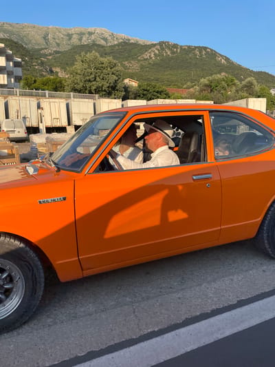 In Zaljevo, Montenegro, an orange vintage Toyota Corolla is parked on a paved road. The car's driver, a man wearing a white shirt and a white fedora, is seated behind the steering wheel. Next to him, a woman in a white top is also in the front passenger seat. In the back seat, a young child with dark hair is visible. The car is positioned in front of a row of large, cream-colored shipping containers. Beyond the containers, a multi-story building and a cluster of trees are situated at the base of a rugged, tree-covered mountain range. The sky above is a clear, bright blue, and the late afternoon sun casts long shadows from the car and surrounding objects onto the road.