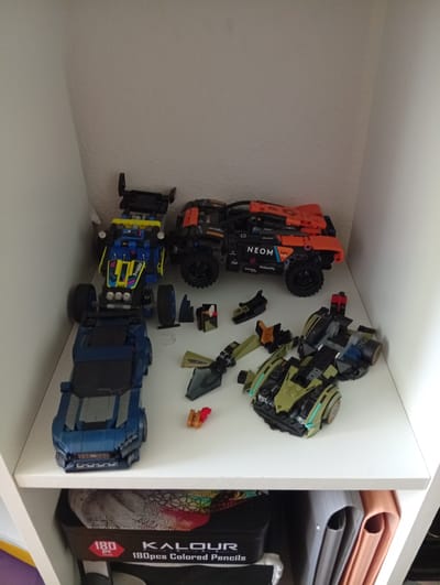 The image shows several toy vehicles assembled from building blocks, placed on a white shelf. In the background, the wall appears to be textured. In the foreground, there is a black box containing colored pencils, with the brand name "KALOUR" and "180 pcs Colored Pencils" visible in white text.

The toys are arranged on the shelf in a playful manner. On the left, a blue car is positioned below a yellow and blue buggy. To the right of the buggy is an orange and black off-road vehicle. In the lower right corner, a green and black toy vehicle, possibly a futuristic car, is also visible. Scattered around the toys are individual building block pieces, some of which appear to be weapon-like accessories.

The scene appears to be indoors, likely in a child's room or a display shelf. The lighting is neutral, suggesting it's daytime, but no specific time is discernible. There are no people or animals in the image. The overall impression is one of organized play and display of assembled toys.

The visible text on the pencil box reads "180 pc" within a red hexagonal shape, followed by "KALOUR" and "180pcs Colored Pencils". There is also some text on the orange and black vehicle, including the word "NEOM" in large letters. The yellow and blue buggy has "buggy" written on its side.