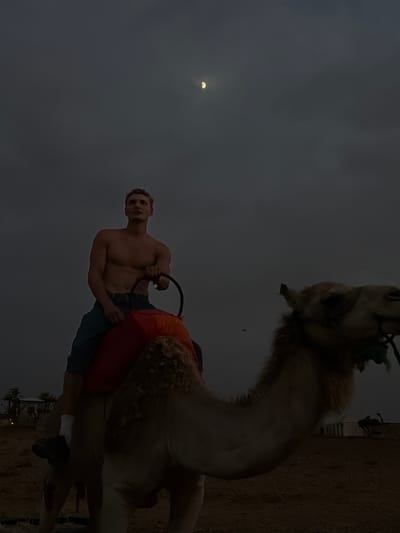 A man with a muscular physique, shirtless and wearing dark shorts and white socks, is seated on a camel. The camel is adorned with a red saddle pad. The man holds onto a dark ring-shaped rein. The scene takes place in Douar Ait Daoud, Morocco, under a dark, cloudy sky with a sliver of moon visible. The ground appears to be sandy or dusty. In the background, there are tents and silhouettes of palm trees, suggesting a desert or oasis setting.