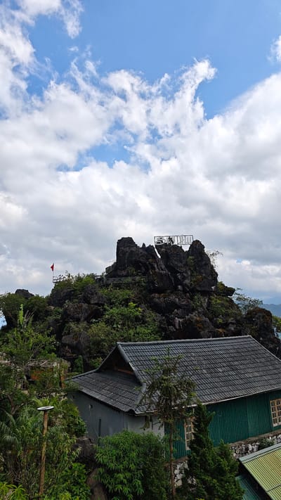 The image shows a view in Tả Van, Vietnam. In the background, a rocky hill with green vegetation is topped by a sign. A red flag is also visible on the hill. A building with a grey tiled roof and green walls is in the foreground. The sky is blue with white clouds. Green trees and bushes surround the building.