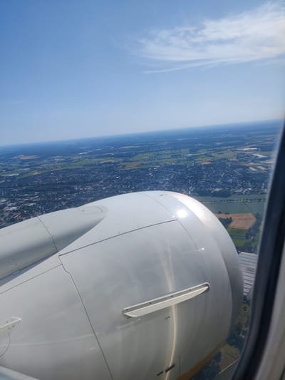 This media file captures an aerial view from an airplane window, looking out over the town of Kevelaer, Germany. The primary subjects are the landscape below and the wing of the aircraft in the foreground. The scene is a daytime view, with clear blue skies and scattered wispy clouds, suggesting pleasant weather.

The airplane's engine cowling is prominently featured, a large, smooth, white object that dominates the lower portion of the image. It reflects the bright sunlight, creating a glare. The window frame is visible on the right side of the image, framing the view of the ground.

Below, a dense residential area of Kevelaer is visible, characterized by numerous buildings with dark roofs. This urban sprawl transitions into patchwork fields and green spaces, with some trees and what appear to be greenhouses or agricultural structures in the distance. The overall impression is of a landscape viewed from a significant altitude during a flight.

There are no people visible in the media file. The activity taking place is implied to be air travel. Notable details include the bright sunlight indicating it is likely midday. The location is specifically Kevelaer, Germany. No discernible text is visible on the engine cowling, though there are small markings that are too small to read. The overall emotion evoked is one of travel and observation.