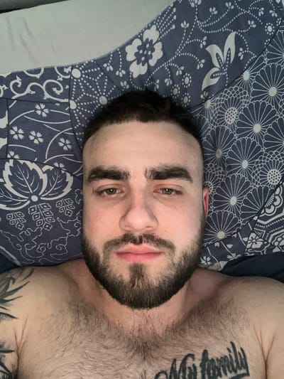 A close-up, overhead shot shows a man lying on a bed. The man has a beard and short dark hair. He is wearing a blue and white patterned shirt. The bed appears to be in a bedroom. The scene is indoors.