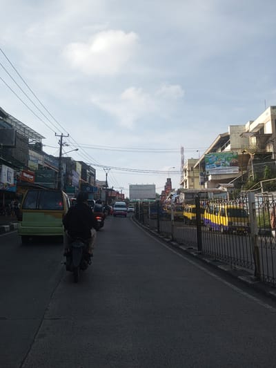 The image depicts a street scene with vehicular traffic in what appears to be a bustling urban area. The location is not Phou Khoun, Laos, as evidenced by the signage and vehicles which are more consistent with urban Southeast Asia. A motorcyclist is in the foreground, followed by a light-green van. Several other vehicles, including a white ambulance, are further down the street. Buildings flank both sides of the road, with various businesses displaying signage. A metal fence runs along the right side of the street. Overhead power lines are visible against the sky. The sky is light blue with scattered clouds. The lighting suggests it's daytime.
