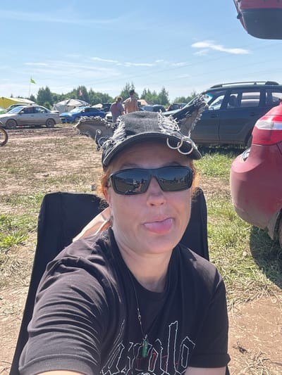 In the city of Chormoz, Russia, a person with reddish hair and wearing sunglasses and a black cap with distressed fabric and rings is sitting in a black chair. They are sticking their tongue out at the camera. The person is wearing a black t-shirt with a pendant necklace. In the background, there are several cars parked on a dirt and grass field, with trees and tents visible in the distance under a blue sky with some clouds. The scene appears to be a festival or outdoor gathering.