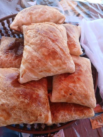 The image shows a close-up, overhead view of a pile of freshly baked samsa, a savory pastry popular in Central Asia, displayed in a woven basket. The samsa are golden brown and flaky, with visible layers of dough. A white cheesecloth is draped to the right of the basket. The scene appears to be an outdoor market stall, possibly in Gulistan, Uzbekistan, given the context of samsa as a local specialty. The background is blurred, but suggests a colorful, possibly patterned tablecloth. There are no people visible in the frame.