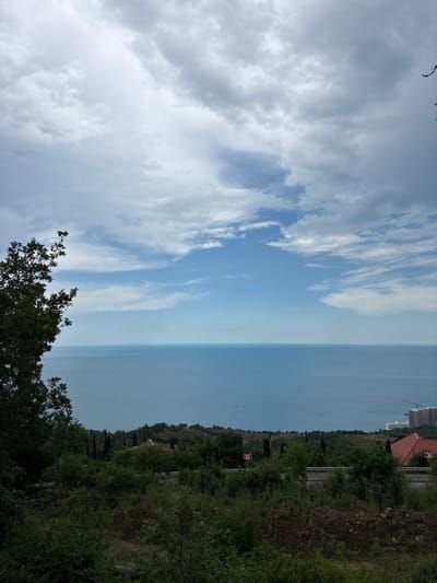 The image depicts a panoramic view of the sea and sky from a hillside, possibly near Malyi Mayak, Ukraine. In the foreground, lush green vegetation and trees dominate the scene. A paved road with a metal railing runs horizontally across the mid-ground. Beyond the road, the landscape transitions to rolling hills covered with trees and some buildings, including a red-tiled roof visible on the right. In the far distance, the vast expanse of the calm, blue sea meets the horizon. The sky above is filled with a mix of light and dark clouds, with patches of blue sky visible in between. A construction crane and several white buildings are visible on the distant coastline. The overall impression is one of a natural landscape with signs of human development.