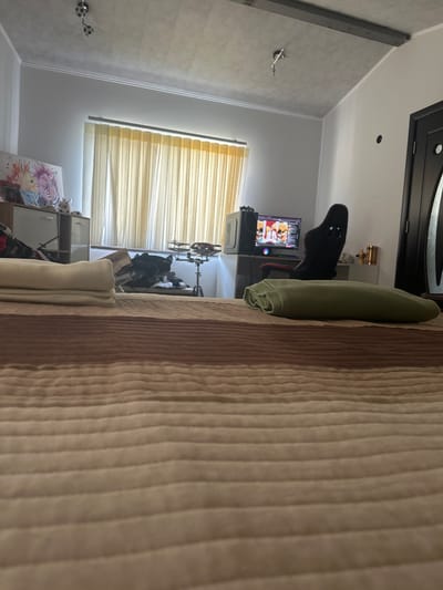 The image shows a room in Madara, Bulgaria. The scene appears to be a bedroom or living area, with a bed or sofa dominating the right side of the frame, covered in a textured fabric with vertical stripes in shades of beige and light brown.

On the left side of the image, a wall is visible with light grey, horizontally paneled wallpaper. A dark grey beam runs vertically along the wall. A wall-mounted object with two spherical decorative elements, possibly knobs or hooks, is attached to the wall. Further down the wall, a window with horizontal blinds, likely made of a light-colored fabric, is partially visible.

In the background, a gaming chair with a black frame and red accents is positioned in front of a television screen which displays an image of people. A computer tower is visible beneath the TV, and a shelf unit containing some items is also present. A light green rolled-up towel or blanket is placed on the bed or sofa. The overall lighting suggests it might be daytime, though the specific time is not discernible. No emotions are explicitly conveyed. There is no visible text in the image.