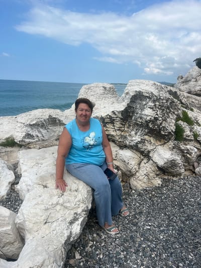 A woman is sitting on a large, white, rocky outcrop near the sea. She is wearing a blue t-shirt with a graphic on it, and blue jeans. Her feet are clad in sandals. She is holding a black smartphone in her left hand. The background shows the blue sea and a cloudy sky. The rocky area is a mix of large white rocks and smaller dark pebbles. The weather appears to be clear and sunny. The woman is looking at the camera with a slight smile. The scene evokes a sense of relaxation and enjoying nature. The location is Tsandrypsh, Abkhazia. The time of day appears to be daytime.