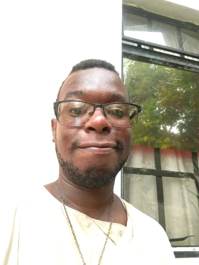 The image shows a Black male, with a short haircut, wearing black-rimmed glasses, a white shirt and a gold chain necklace, looking directly at the camera. He has a 5 o'clock shadow. He is standing in front of a white wall and a window with black trim. The window is covered in a white curtain. The window is reflecting trees outside. The scene is located at coordinates 10.248, -61.483, which are near the location of La Romaine, Trinidad and Tobago.