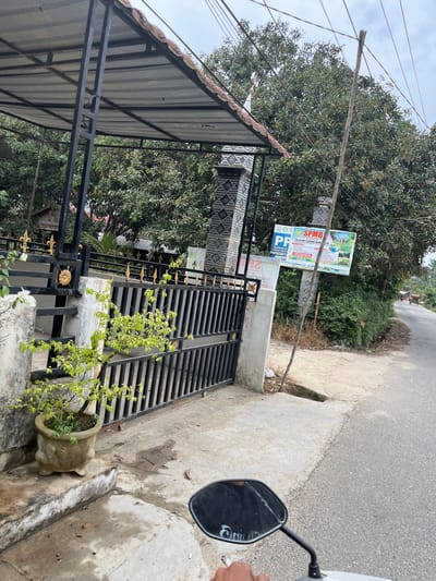 The image depicts a street-level view of a residential area or school entrance in Tanahterban, Indonesia, captured from the perspective of someone riding a motorcycle. In the foreground, the handlebar and a portion of a white motorcycle are visible, along with a black rearview mirror reflecting the sky and trees. A hand holding a cigarette is also present.
The focal point of the scene is a black metal gate with decorative gold accents, opening onto a concrete pathway. To the left of the gate, a potted plant with vibrant green leaves sits on a weathered concrete step. The gate is set against a white concrete pillar adorned with patterned tiles.
Behind the gate, lush green trees dominate the background, some bearing visible fruit that appears to be rambutan. A large sign with Indonesian text and colorful graphics, likely advertising a school or educational program, is affixed to a wooden pole amidst the foliage. The sign's text includes "SPMB TK ISLAM TERPADU ALAM" and "PENERIMAAN TAHUN PELAJAR".
The street itself is paved and appears to be a two-lane road with a slight incline. A few distant figures on bicycles or motorcycles can be seen further down the road, suggesting a moderate level of activity. The sky is overcast, contributing to the overall subdued lighting of the scene. The environment appears to be rural or semi-urban, characterized by dense vegetation and modest structures.