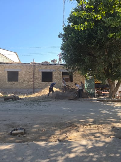 The image depicts a construction site in the city of Shiklar, Uzbekistan, on a clear, sunny day. A partially constructed building with brick walls and pre-fabricated concrete roofing panels is visible. Several men are engaged in construction work. One man is digging a pile of dirt with a shovel. Others are standing around the site, with one man operating what appears to be a cement mixer on the roof. A large tree with green foliage is prominent in the foreground on the right side of the image. The ground is covered in dirt and debris, indicating ongoing construction activity. Power lines are visible against the blue sky.