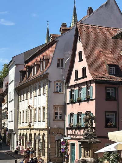 The image depicts a street scene in Bamberg, Germany, likely in the historic center given the architecture. In the foreground, people are visible walking and milling about on a cobblestone street. To the right, a cream-colored umbrella shades an outdoor seating area. Dominating the middle ground are several multi-story buildings with characteristic Bavarian tiled roofs and ornate facades. One building is painted a pale pink and features blue shutters on its windows. A stone fountain topped with a sculpture of an eagle stands prominently in front of this pink building. Other buildings are lighter in color, some with multiple arched windows and decorative stonework. In the background, church spires with crosses rise above the rooftops, indicating a religious presence in the vicinity. Trees are visible on the left side of the street, suggesting a blend of urban and natural elements. The sky is clear and blue, indicative of fair weather. The overall impression is that of a bustling, charming European city street on a sunny day.