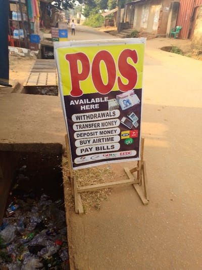This image depicts a street scene in Agukwu Nri, Nigeria, under bright, sunny conditions, likely during the morning or late afternoon given the warm light. The main subject is a brightly colored Point of Sale (POS) service sign, prominently featuring large red "POS" lettering on a yellow background, supported by a simple wooden stand. The sign advertises "AVAILABLE HERE" services including "WITHDRAWALS," "TRANSFER MONEY," "DEPOSIT MONEY," "BUY AIRTIME," and "PAY BILLS." It also displays logos of Nigerian telecommunications companies (MTN, Airtel, GLO) and other service providers (StarTimes, GOtv, EEDC), alongside images of Nigerian Naira banknotes and a POS terminal.
The sign stands beside an open concrete drainage ditch on the left, which is heavily cluttered with plastic bottles, bags, and various trash items. A dirt road stretches into the background, where a lone, blurry figure of a person can be seen walking. Simple, utilitarian buildings, some with corrugated metal details, line both sides of the road in the distance. Further along the drainage ditch, a wooden grate acts as a makeshift bridge. The immediate ground around the sign is bare earth with sparse dry grass. The scene contrasts the modern financial services advertised with the informal and somewhat unkempt setting of the street.