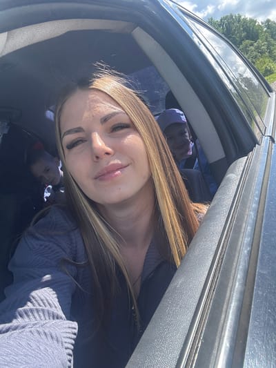 A woman with long, light brown hair, wearing a light blue sweater, is positioned in the front passenger seat of a dark-colored car. She is looking at the camera with a slight smile. Through the car window, vegetation and a partially visible sky suggest an outdoor setting. There are two other people are also visible in the reflection of the car window. Given the angle and the framing, the image appears to be a self-portrait taken by the woman. The location in Slobodka, Russia cannot be determined from the image.