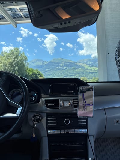 The image shows the interior of a car, likely a Mercedes-Benz, parked under a structure with solar panels, with a scenic mountain landscape visible through the windshield. The car's dashboard is prominent, featuring a steering wheel, instrument cluster, central infotainment screen, and navigation system displayed on a smartphone mounted to the dashboard. The navigation app shows a route with "Raststätte Rheintal" as a destination. The dashboard also includes various buttons and controls for the car's functions, as well as a digital clock and an analog clock. Keys are in the ignition.
The outdoor environment is a bright, sunny day with fluffy white clouds against a blue sky. Lush green trees and rolling mountains form the backdrop. The presence of "Raststätte Rheintal" on the navigation suggests the car is either approaching or has passed this rest stop, which is located on the A13 motorway in the Rhine Valley in the canton of St. Gallen, Switzerland. Buchs (SG), Switzerland, is a town in the Rhine Valley and is geographically close to this area, making it a plausible general location. The overall scene depicts a moment of travel or a stop during a road trip in a mountainous region.