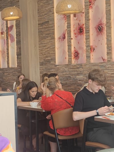 The image depicts an indoor dining area, possibly a restaurant, with a noticeable theme of floral decor. The walls are constructed from stacked, irregular brown stones, and large vertical panels featuring pale pink floral designs, resembling cherry blossoms, are hung between sections of the stone wall. Overhead, woven basket-like light fixtures hang, casting a warm glow.
In the foreground, several individuals are seated at tables. On the right, a young man with curly brown hair, wearing a black polo shirt and dark pants, is looking down at his phone. He has a watch on his left wrist. In front of him is a plate with pasta. To his left, a woman in a vibrant red top, her back mostly to the camera, is also seated. A dark handbag is slung over her shoulder.
Further to the left, a woman with long dark hair, wearing a dark brown top, is looking at her phone and holding a white bowl. She is wearing a bracelet on her left wrist. Beside her, another woman with long brown hair is partially visible. In the background, more people are seated at tables, and the floral wall panels are prominent. The overall impression is a lively dining setting, likely a restaurant with an Asian-inspired aesthetic given the floral motifs. The specific location of Sint-Pieters-Leeuw, Belgium is not visually indicated in the image itself.