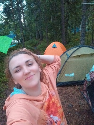 A woman with curly auburn hair, winks and smiles as she poses for a selfie outdoors. She is wearing an orange hoodie with a colorful print on it. Behind her, a light green tent with the word "KOLO2" printed on it is visible. Further in the background, an orange tent is also present, along with other tents that are partially visible in the distance. The scene is set in a forest, with trees and foliage surrounding the campsite. The ground is covered in dirt and dry leaves, suggesting a natural, wooded environment. The overall lighting indicates it is daytime, possibly late afternoon or early evening. The location is identified as Tarasovskoe, Russia.