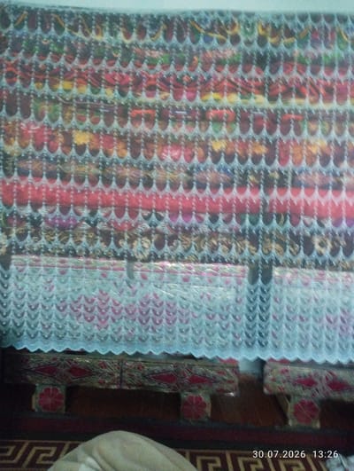 The image shows a decorative textile, likely a curtain or a runner, with an intricate pattern of interwoven threads in various colors including red, pink, yellow, green, and blue. The textile features a repeating geometric design of scalloped edges. Below the textile, there appear to be two upholstered seating structures, possibly low benches or ottomans, with a patterned fabric that includes red diamond shapes and a circular floral motif in red. The flooring is covered by a dark carpet with a geometric Greek key pattern. The lighting suggests an indoor setting. The timestamp "30.07.2026 13:26" is overlaid on the bottom right of the image, but this does not indicate the actual location of the scene in Quyosh, Uzbekistan.