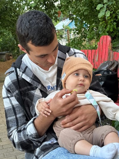 A man is holding a baby outdoors in a natural setting. The man has short, dark hair and is wearing a white t-shirt under a black and white plaid shirt. The baby is wearing a light brown beanie and a beige knit sweater, and brown waffle-knit pants. The baby has bright blue eyes and is looking towards the camera. The man is holding a small, yellow object near the baby's mouth, possibly food or a toy.

The scene appears to be a park or garden, with lush green foliage and trees in the background. A red, weathered wooden structure is visible behind the man and baby. The ground beneath them appears to be paved with light-colored bricks or stones. A black bag rests on the red structure. The lighting suggests it is daytime, possibly overcast as there are no strong shadows. The overall mood is one of tenderness and care, as the man is gently holding and interacting with the baby. A tag on the baby's beanie has text that is partially visible, but not clearly legible.