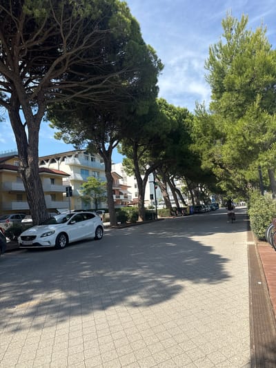 The image shows a tree-lined street in Lignano Sabbiadoro, Italy, on a sunny day. A white Volvo V40 is parked on the left side of the street, with its license plate reading "KLE446EW". A woman is cycling on the right side of the street, and there are several buildings and trees visible in the background. The street is paved with light-colored tiles, and shadows from the trees are cast across the pavement. In the distance, more parked cars and a few people can be seen. The overall scene depicts a tranquil urban environment with abundant greenery.