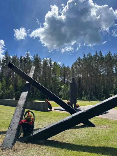 The image depicts a memorial site located in Акалоўскі сельскі Савет, Belarus. In the foreground, two large, dark, rough-hewn beams are crossed and placed on the ground, forming an "X" shape. To the left of the crossed beams, a black basket filled with red flowers rests on the grass. In the mid-ground, a somber, dark-colored statue of a person stands facing away from the camera, with a wreath of red flowers at its base. Behind the statue, a low concrete wall stretches across the scene. Further in the background, a dense forest of tall pine trees forms a natural backdrop. The sky above is bright blue, with scattered white clouds, indicating a sunny day. The ground is covered in green grass. The overall impression is one of solemnity and remembrance.