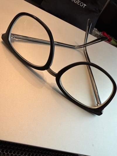 The image shows a pair of black-rimmed eyeglasses with clear lenses resting on a light-colored, textured surface. The eyeglasses are positioned diagonally across the frame, with the left lens closer to the viewer and slightly angled upwards. The right lens is positioned further back and is more oriented towards the viewer. The metal temples of the glasses are visible, crossing over each other. In the background, a dark object with reflective text that appears to say "PEUGEOT" is partially visible, along with some colorful items. The lighting suggests an indoor environment. The location in the city of Biržai, Lithuania is not determinable from the visual information provided in the image.