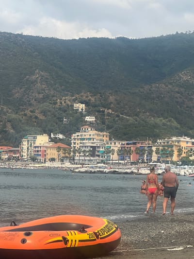 The image depicts a panoramic view of a coastal town, identified as Sestri Levante, Italy, during daylight hours, observed from a low vantage point on a pebble beach.
In the immediate foreground, an orange and black inflatable boat, an "INTEX 200 EXPLORER," rests on the wet, dark grey pebble beach, with its bow extending slightly into the shallow, rippled water. To the right of the boat, a woman in a red bikini and a man in dark swim shorts are walking away from the viewer, entering the water.
The midground features a calm, dark blue-grey body of water. Several individuals are visible in the water, some partially submerged. A line of grey rock breakwaters extends across the water, separating the inner beach area from the more open bay. Beyond the breakwaters, numerous white beach umbrellas and sun loungers are densely arranged on a section of the beach, indicating a populated recreational area. Small boats are moored in the water behind the breakwaters.
The background is dominated by the town's architecture and the surrounding natural landscape. Multi-story buildings, characteristic of Mediterranean coastal towns, line the shoreline. These structures display a palette of colors including yellow, pink, orange, light blue, and white, with visible balconies and awnings. Palm trees are interspersed among the buildings. Directly behind the town, steep, densely vegetated mountains rise sharply, covered in green foliage. Several individual houses, including a prominent light-colored structure, are situated high on the mountain slopes. The sky above is overcast with scattered grey clouds.