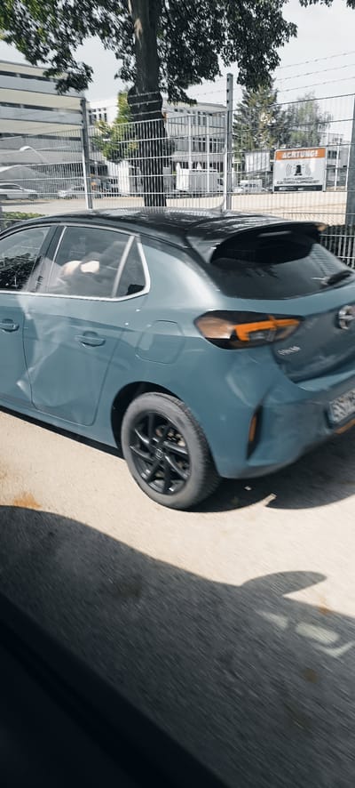 The image shows the rear passenger side of a blue Opel Corsa hatchback driving past a fence. The car has a black roof and black alloy wheels. The car's rear lights are visible, with amber turn signals illuminated. Inside the car, two people are visible in the rear seats, appearing to be looking towards the driver.
Behind the car and fence, a white building with multiple windows is visible. A sign with "ACHTUNG!" and a siren symbol is affixed to the fence. The ground is asphalt.
The image was taken in Fellbach, Germany, specifically near the border with Stuttgart, along the Bundesstraße 14. The time of day appears to be daytime, with sunlight casting shadows on the ground.