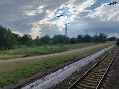 The image shows a view of a railway track and its surroundings in Novokubansk, Russia. In the foreground, a train track runs diagonally from the lower right to the upper left of the frame. The track is bordered by a low, white concrete barrier, which shows signs of wear and peeling paint. Beyond the track, a dirt path runs parallel to it, separating the track area from a field of green grass and vegetation. In the middle ground, a line of trees and bushes forms a dense green barrier. In the background, several tall electrical pylons with overhead wires are visible, indicating the presence of a power transmission line. The sky is partly cloudy with patches of blue sky and sunlight breaking through the clouds. A white vehicle is visible in the distance on the dirt path, near the trees. The scene appears to be a rural or semi-rural area within the city, likely along the outskirts where infrastructure like railways and power lines are common.