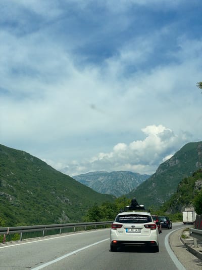 The image depicts a mountain road scene, likely in a rural area. A white car, identifiable as an Apple Maps Imagery Collection vehicle based on the sticker on the back, is driving on the road. A person is seen behind the car. A truck is visible further ahead. The road winds through a mountainous landscape, with green hills and cliffs on one side and a partly cloudy sky above. The location is potentially in Donji Jasenjani, Bosnia and Herzegovina, inferred from the Apple Maps vehicle's data collection. The perspective is from inside a car.