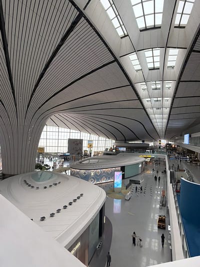 This image captures a wide-angle view of a modern airport terminal in Guangyang District, China. The architecture is characterized by a sweeping, shell-like roof structure with a repeating ribbed pattern, supported by large, curved columns. A significant portion of the ceiling is made of skylights, allowing natural light to illuminate the spacious interior.

On the ground level, there is a large, open concourse area with polished floors that reflect the overhead lighting. Several shops are visible, including one with prominent "NCIAGA" signage, likely part of a fashion retailer. In the center of the concourse, there is a circular kiosk or display area with a patterned facade. People are dispersed throughout the terminal, some walking, some pushing luggage carts, and others standing. A yellow baggage cart is parked near the right side of the frame. The overall environment suggests a busy but organized airport setting.