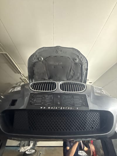 This is a low-angle, close-up, indoor, eye-level shot of the front of a dark gray car with its hood open, angled upwards, in what appears to be a garage or workshop in Čelákovice, Czechia. The car's grille, headlights, and the underside of its hood are visible. A license plate frame with the text "www.majagercy.cz" is mounted on the front bumper. Below the bumper, a black mesh grille is visible, and further down, a tire and a white cloth are on the floor. The ceiling, illuminated by fluorescent lights, is visible in the upper portion of the image.