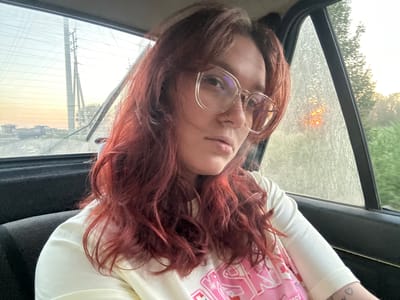 The image depicts a young woman with reddish-brown hair, wearing large clear-rimmed glasses and a white t-shirt with a pink graphic, as she sits in the passenger seat of a vehicle. She is looking towards the camera with a neutral expression. Through the rear window of the car, power lines and telephone poles are visible against a hazy sky, suggesting a rural or semi-rural setting. The side window shows foliage and a blurred orange glow, possibly the sun setting or a street light. There is a faint sheen of dirt or dust on the rear window. The interior of the car is visible, with dark seats and a portion of the door panel. The image appears to have been taken during the daytime, likely in the late afternoon or early evening due to the lighting conditions. The specific location of Ленин, Kyrgyzstan is not discernible from the visual cues in the image.