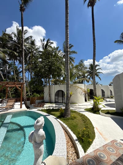 The image shows a tropical scene in Gubug, Indonesia. Palm trees stretch across a bright blue sky dotted with white clouds. The lower portion of the image features white buildings with arched windows and greenery. A pool with a statue is visible, bordered by a white tile walkway and a patterned tile area. A wooden structure sits near the pool.