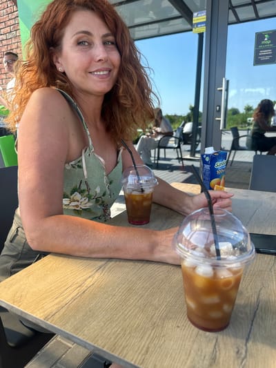 A medium-shot, eye-level outdoor photo shows a woman sitting at a wooden table in front of a building with a glass facade. She has auburn, curly hair and is wearing a green top with a floral pattern and khaki pants. Her left arm is resting on the table, and she is looking at the camera with a smile. Two iced coffee drinks with straws are on the table, along with a carton of orange juice. In the background, other people are seated at tables, and trees and buildings are visible outside. The scene appears to be a cafe or restaurant in Syrnyky, Ukraine.