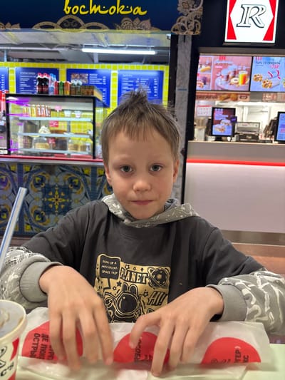 A young boy is sitting at a table in a fast food restaurant. He has light brown hair and blue eyes. He is wearing a dark gray hooded sweatshirt with a graphic print of an astronaut. He is holding a wrapped food item in his hands. The restaurant has blue and yellow decor. The restaurant name "bochmokia" is visible on the left side. In the background, a menu board with images of food and prices is visible, along with other customers and employees. The words "DIARY OF A MYSTERIOUS SPACE TRIP" and "PLANET" are visible on the boy's sweatshirt. The food item has red circles printed on its wrapping. The text "DE LOKE OSTRY" and "SOBBI" are visible on the wrapping. Based on the Cyrillic text and other signs, the location is likely in Grushovaya Balka, Russia.