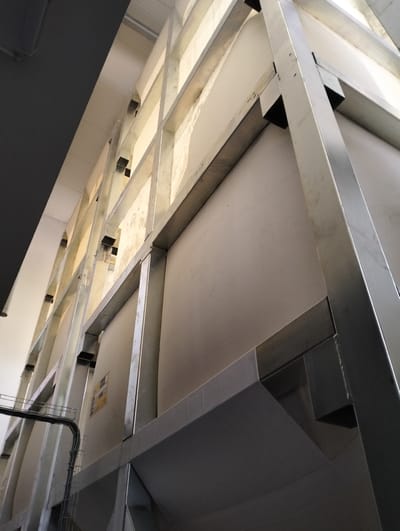 The image depicts the interior of a large industrial structure, likely a factory or processing plant, in Șoldănești, Moldova. The dominant elements are a complex framework of metallic beams, likely aluminum or steel, forming a grid-like support system. These beams are arranged both vertically and horizontally, creating a sense of depth and scale.

Attached to this metal framework are large, flat panels, appearing to be made of a light-colored, matte material, possibly some form of insulation or structural paneling. These panels cover large sections of the interior, creating a segmented, wall-like appearance within the structure. The lighting suggests natural light is filtering in from above or from windows or openings not fully visible in the frame, casting shadows and highlighting the metallic textures and the surfaces of the panels.

A dark, angular structure, potentially a duct or conduit system, is visible in the lower left portion of the image, suggesting the presence of utilities or a production line. A yellow warning or informational label is affixed to one of the large panels in the middle ground, adding a detail of functionality and regulation to the industrial setting. The perspective is from a low angle, looking upwards, emphasizing the height and industrial scale of the facility. No people are visible in the image.