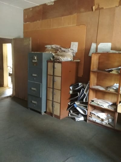 The image depicts an indoor scene, likely an office or storage area, with a grey metal filing cabinet and a wooden shelving unit as prominent objects. The filing cabinet, on the left, has four drawers. Next to it is a narrower, taller filing cabinet with lighter-colored drawers. To the right is a wooden bookshelf with several shelves, containing stacks of papers, folders, and binders. Papers are also piled on top of the filing cabinet and on a nearby surface. The walls appear to be paneled with a warm-toned material. The floor is covered with a dark, textured carpet. A doorway is visible on the left, leading to another room. The scene is cluttered with documents, suggesting it might be an archive or a space with unmanaged paperwork. There are no individuals or active interactions visible in the image. The location is identified as Bisichi, Nigeria.