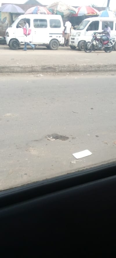 The image is a view from a car window looking out onto a street in Igrita, Nigeria. There are a few white vans parked along the side of the road. One person is walking away from the camera, and another person is standing near a white van. The road is paved and there is a small patch of dirt in the middle of it. A piece of white paper is lying on the road in front of the patch of dirt. The environment is fairly sparse, with only a few trees and buildings in the background.  The image is taken from a low angle, looking out towards the horizon. The image is slightly blurry, which could be due to the car window being dirty. The image is taken in daylight. The time of day is unknown. The weather is overcast. The image is in focus.