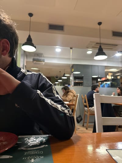 A person sits at a wooden table in a restaurant. A menu card, with the text "PLANTERAY CITADELLE," and a red object, are on the table. The person is wearing a dark blue track jacket with white laurel wreath stripes down the sleeves. Other patrons are visible in the background sitting at tables and chairs. The restaurant has a mirrored wall and pendant lights hanging from the ceiling.