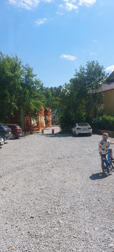 The image depicts a person riding a bicycle on a gravel surface, likely a parking lot or road, in Berdsk, Russia. The person, seen from the rear and side, is wearing a white t-shirt with a colorful pattern and blue shorts, and a light-colored cap. They are on a blue bicycle and are angled towards the right side of the frame.
In the background, a white car is parked. Further back, there are trees with lush green foliage and a building with a tiled roof. Two wooden structures with draped fabric are also visible, possibly gazebos or covered seating areas. The sky is a clear blue with a few white clouds. The scene appears to be set during daylight hours, likely a sunny day, given the shadows and bright lighting.
There is some visible text on the license plate of the white car, which reads "E570XA 194". The overall setting suggests an outdoor recreational area or a residential neighborhood. The activity is simple leisure; someone is enjoying a bicycle ride.