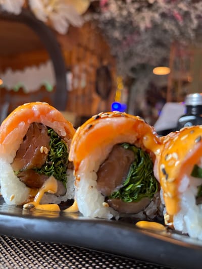 A close-up photograph depicts three sushi rolls arranged on a black rectangular plate, resting on a dark, woven placemat within a restaurant environment in Bad Sobernheim, Germany. Each sushi roll consists of white rice, topped with a slice of orange-pink salmon, glazed with a light orange-brown sauce, and sprinkled with white sesame seeds. The visible interior filling includes dark green leafy greens and a reddish-brown component, likely fish. A creamy, pale orange sauce is drizzled over the rolls and pools on the plate. The background is out of focus, revealing blurred elements of a restaurant interior, including wooden-like architectural arches, various bokeh lights (a distinct blue light and several yellow lights), white floral decor, dark foliage, and a partial view of a dark bottle with a silver cap. No individuals are present in the image.