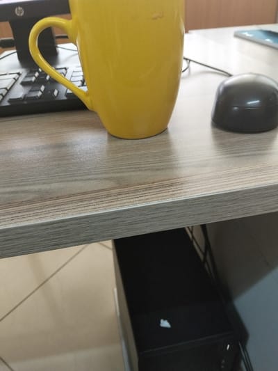 A static, eye-level photograph captures a portion of an indoor workspace in Viwandani ward, Kenya. A yellow ceramic mug, bearing a light-colored mark near its rim, is centered on a light-brown wood-grain laminate desk surface. To the right, a dark gray dome-shaped computer mouse rests on the desk. In the background, partially obscured by the mug, a black computer keyboard is visible on the left, alongside an HP-branded monitor stand. A corner of a white device with a blue screen, likely a tablet or smartphone, is visible on the far right background of the desk. Below the desk, a black computer tower with a small white mark on its top surface is positioned on a light-tiled floor with visible grout lines. No individuals or active interactions are depicted.