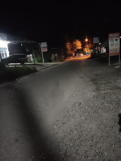 The image depicts a dimly lit street scene at night, likely in the city of Keudematangkuli, Indonesia, given the presence of Indonesian language on a sign. The road surface is unpaved and appears to be a mixture of dirt and gravel. To the left, a dark-colored SUV is parked near a building with lit windows. Further down the road, a street light illuminates the area, casting a yellow glow on the trees and the road. There are several signs visible, one of which appears to be a menu or advertisement with images of food, written in Indonesian. The overall impression is of a quiet, rural or suburban street at night.