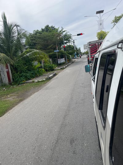 The image shows a street scene in Jurong Peukan, Indonesia, viewed from inside a vehicle. The perspective is tilted, suggesting the photo was taken from a moving vehicle. The road is paved and appears to be an urban or semi-urban environment with greenery on the left side of the road.

Prominent objects include a red traffic light, indicating that vehicles may be stopped. Further down the road, another traffic light is visible, and there are signs that are too blurry to read clearly. A truck with red and black stripes, possibly a delivery truck, is parked or moving on the road. The vehicle from which the photo is taken is white and its side and window are visible.

The setting is characterized by lush green trees and vegetation lining the road. A palm tree is visible in the foreground on the left. Overhead wires and a lamppost are also present. The sky is overcast, suggesting a cloudy day.

The activity taking place appears to be a regular street scene, possibly during daytime traffic, given the traffic lights and the presence of vehicles. No people are clearly visible. The overall impression is that of a typical day in a Southeast Asian town or city. There are some unreadable signs, one of which appears to be a blue street sign.